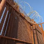 Low angle view of tall metal border wall topped with thick coils of razor wire against a clear blue sky in Naco Arizona.