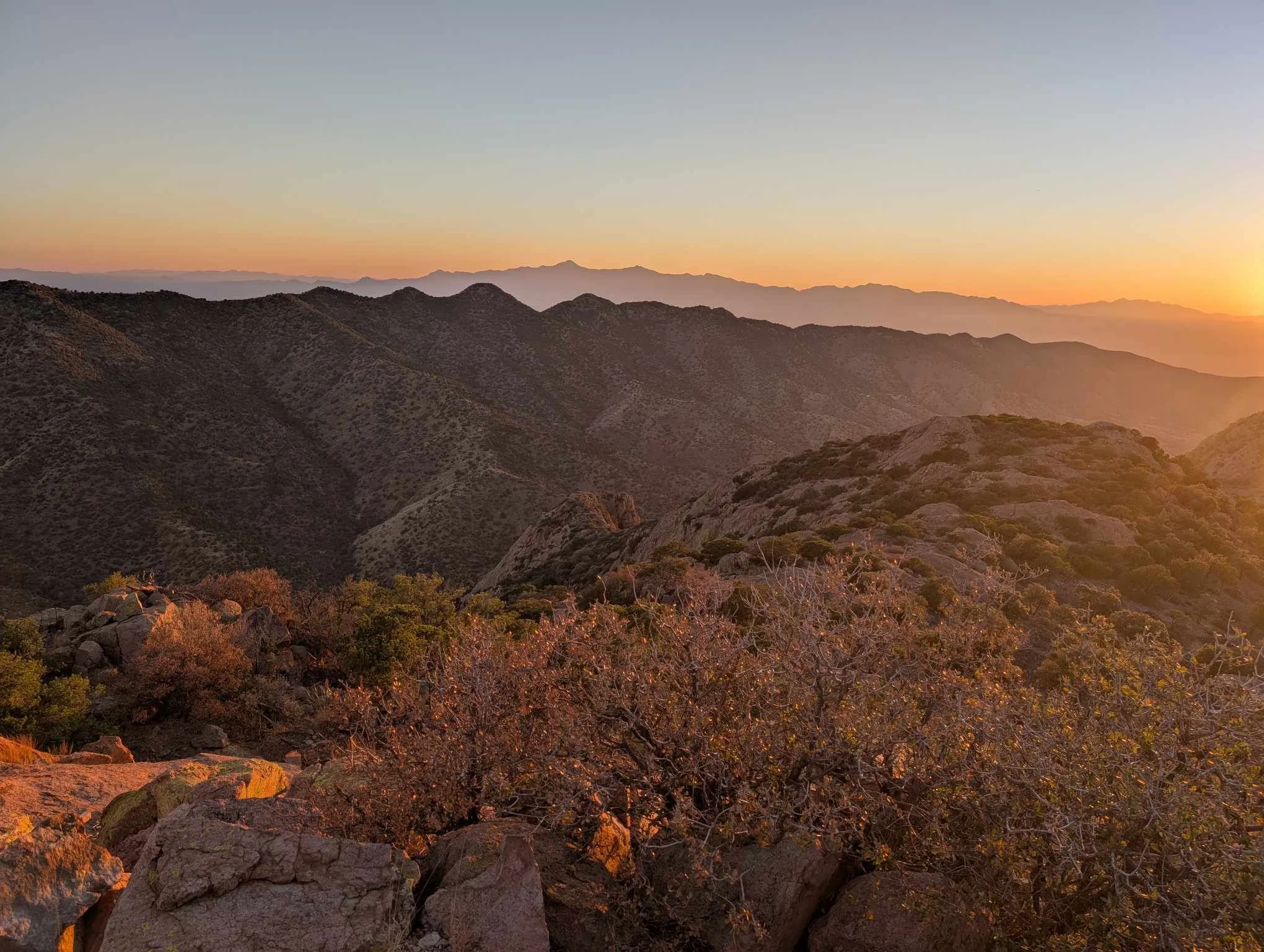 Warm golden hour sunset light illuminating rocky hills and shrubs with layered mountain ranges at Juniper Flats.