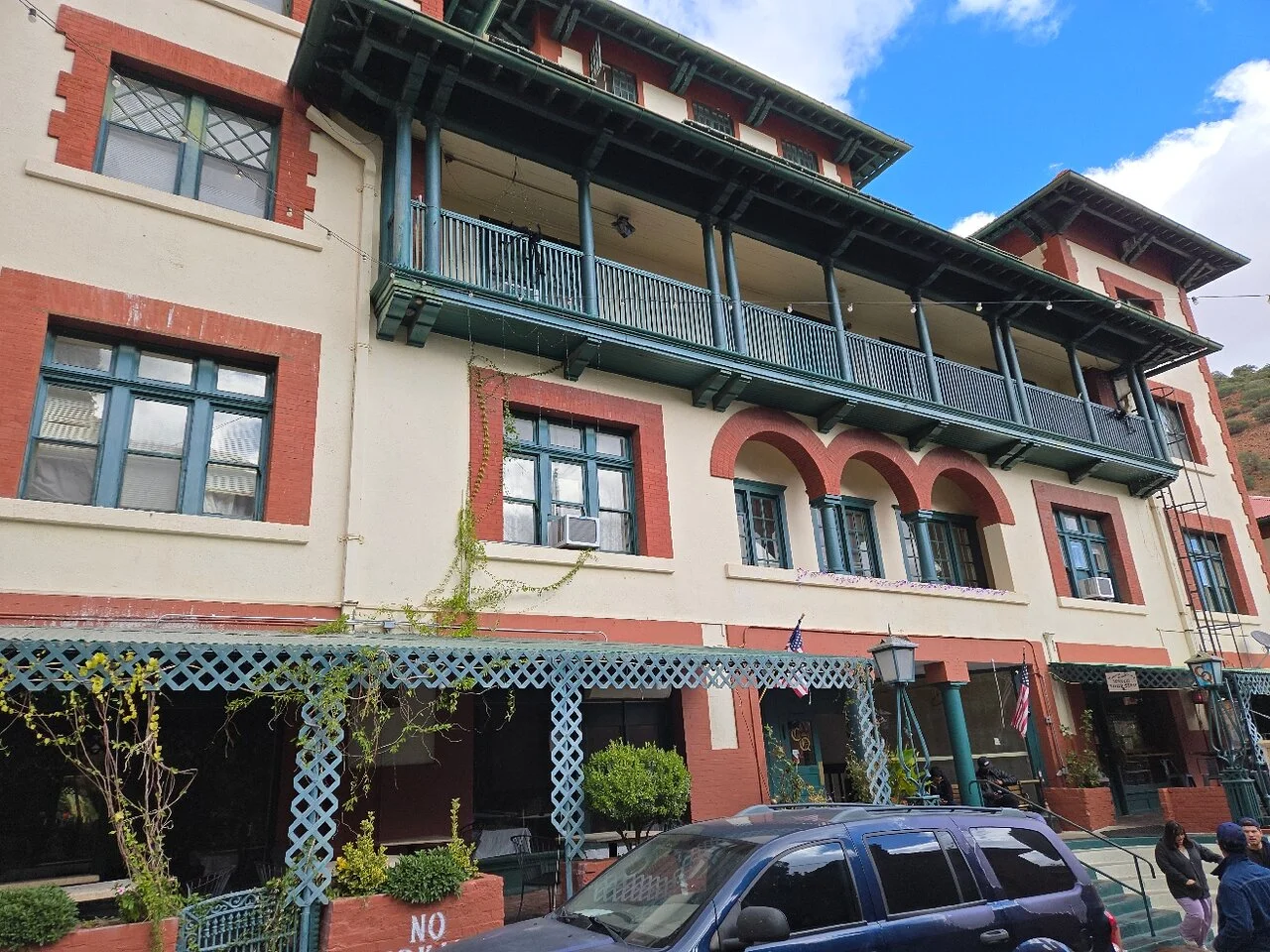 Historic Copper Queen Hotel building with green balconies, red brick trim, and blue SUV parked in front under blue sky.