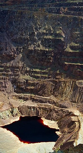 View into Bisbee’s massive Lavender Pit on an overcast day, showing layered mining benches, colorful rock formations, and a dark water pool at the bottom, as seen along the Copper Territory Tour route.