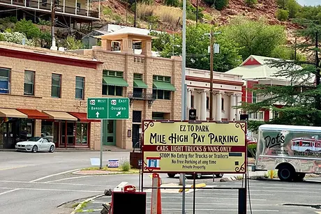 The Mile High Pay Parking lot in historic Old Bisbee, featuring vintage architecture, green highway signs, and a red desert mountain background.