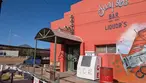 Bright orange Gay 90’s Bar building with airplane mural, ice machine, and Bud Light sign in Naco under blue sky.