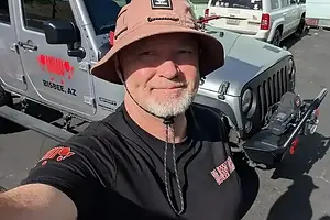 Man wearing bucket hat smiling in selfie with gray Big Jeep Tours vehicle in background.