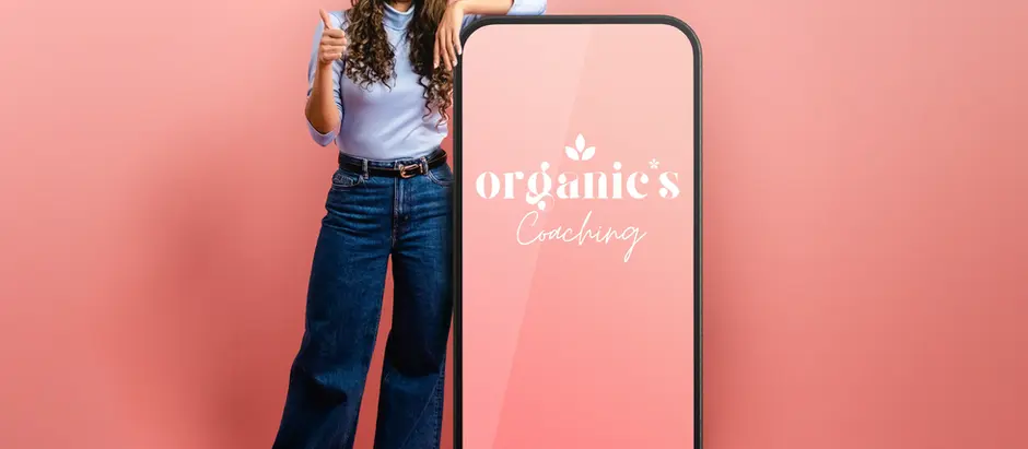 Lächelnde Frau in professionellem Outfit zeigt selbstbewusst auf ein großes Smartphone-Display mit einem eleganten rosa Logo für organics Coaching