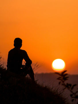 Silhueta de um homem, em um monte com mato baixo, assistindo ao pôr do sol em um horizonte distante, que também se mostra como uma silhueta. A imagem apresenta tons de laranja no céu e preto nas silhuetas.