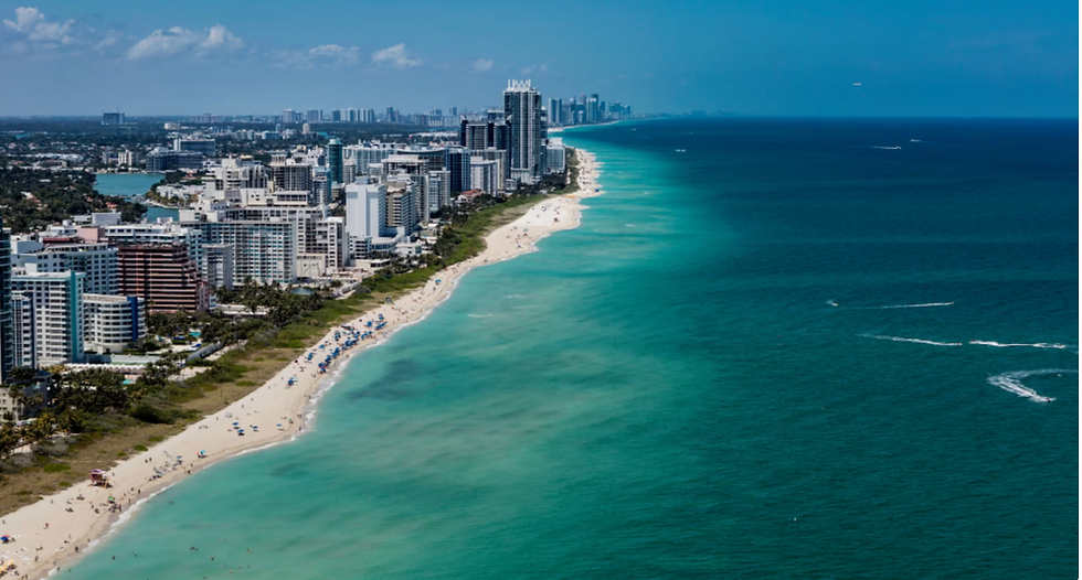 An aerial view of Miami Beach, Florida, showing a long stretch of white sand beach lined with high-rise buildings and turquoise ocean waters extending into the horizon under a clear blue sky.