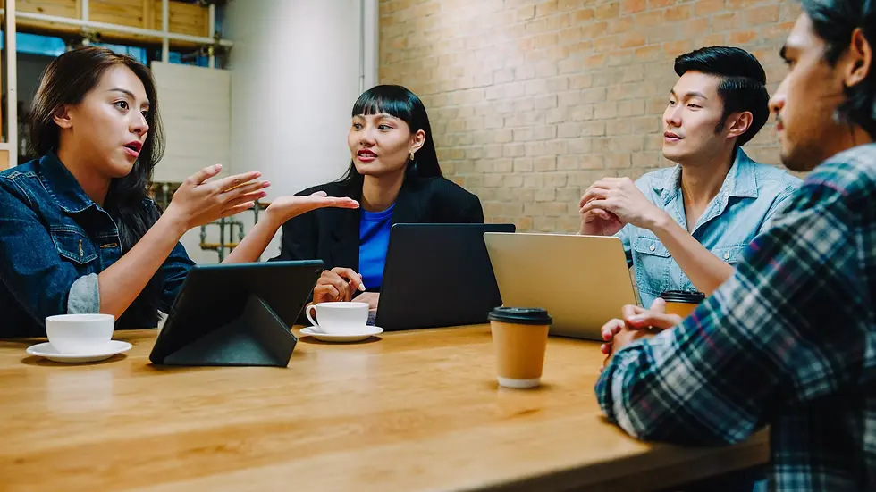 Four people in a meeting room discuss around a wooden table with laptops and coffee. A woman gestures expressively. Brick wall background.