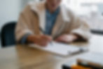 Person in a beige coat writes on a clipboard at a wooden table. Papers and a blurred cityscape window in the background create a focused mood.