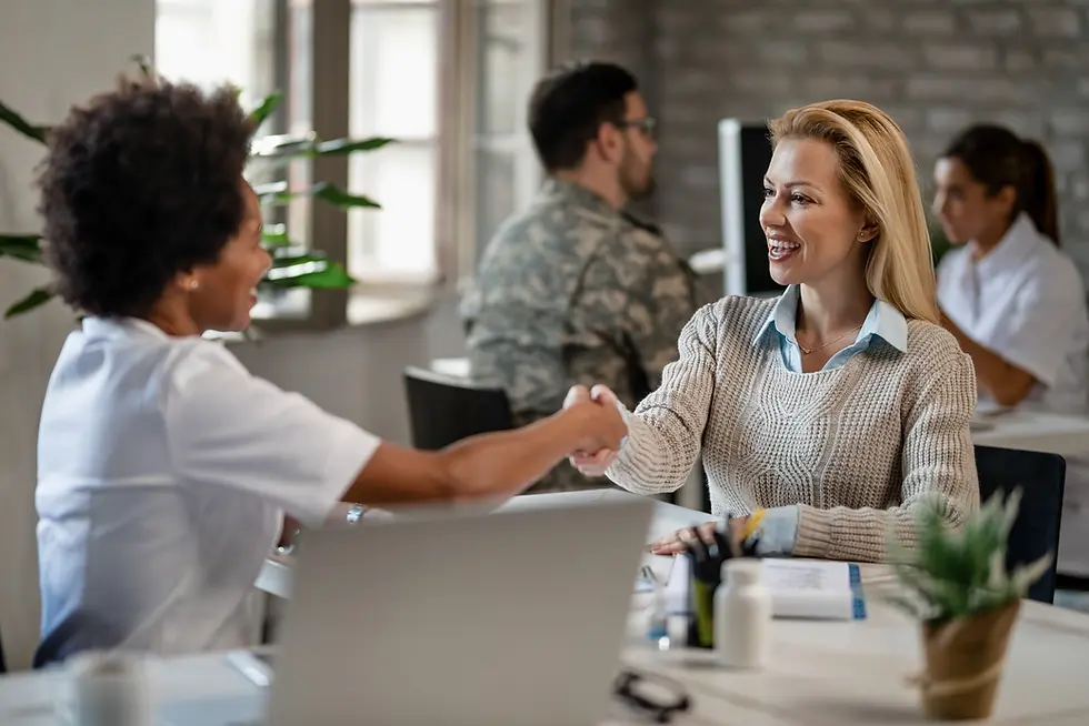 Two women shake hands and smile in an office with laptops. Background shows colleagues working with a plant and soft lighting.