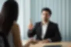 Man in suit raises finger while facing a woman in an office setting with vertical blinds. Serious mood, neutral tones.