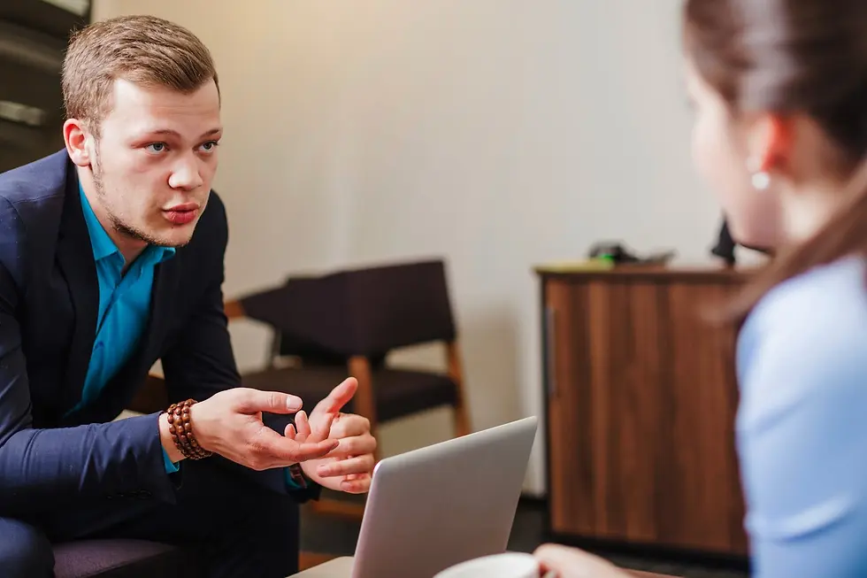 Man in blue suit talking to woman, gesturing hands, in office setting with laptop on table. Engaged discussion, wood cabinet behind.