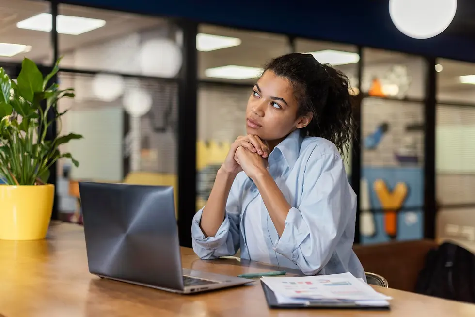 Young woman in an office gazes thoughtfully, hands clasped near chin. Open laptop, papers on table, potted plant in yellow pot nearby.