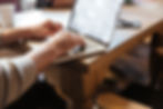 Hands typing on a laptop with visible text on screen. Wooden table surface with a cup and phone nearby. Bright, indoor setting.