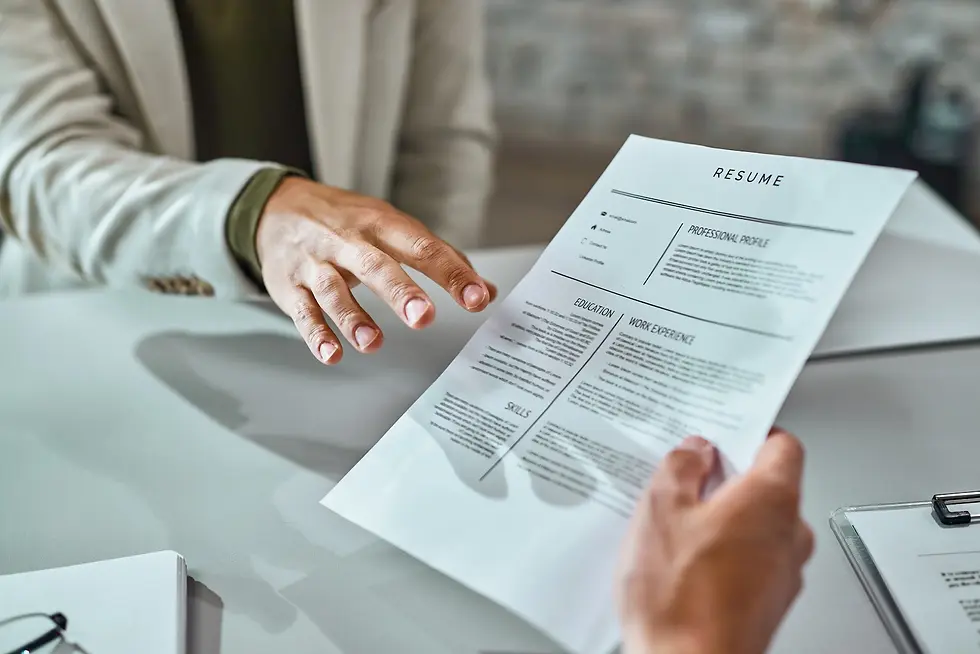 Person handing over a resume across a table in an office setting. Resume shows sections on education and work experience. Professional tone.