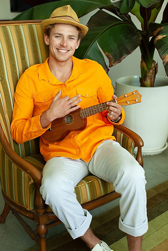 Sustainable Shane plays the ukelele and wears a fedora and yellow shirt in front of a palm.