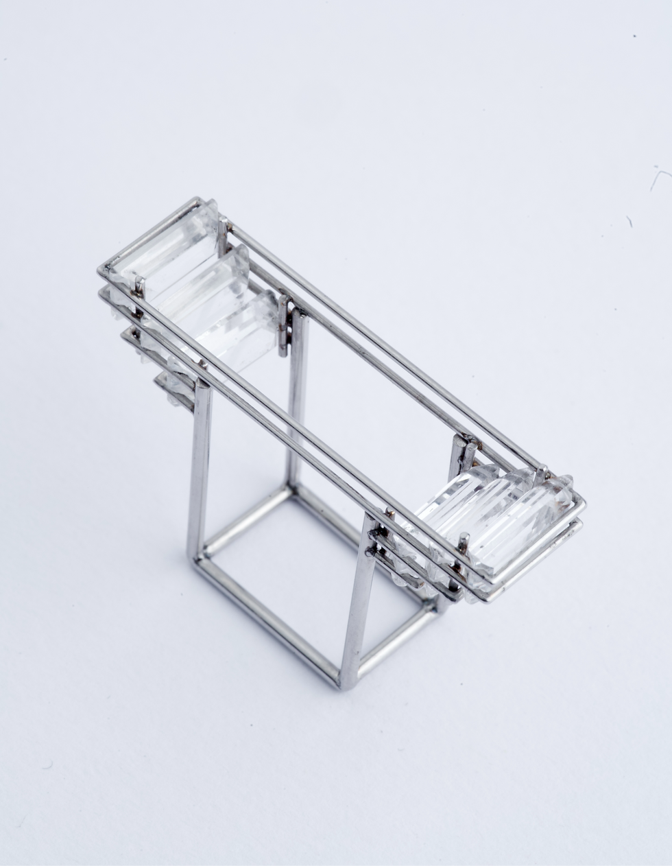 Thumbnail: A square silver ring on a plain white background, photographed from above. The ring been designed to display the finger and showcase the clear gemstones extending from the sides of the ring. 