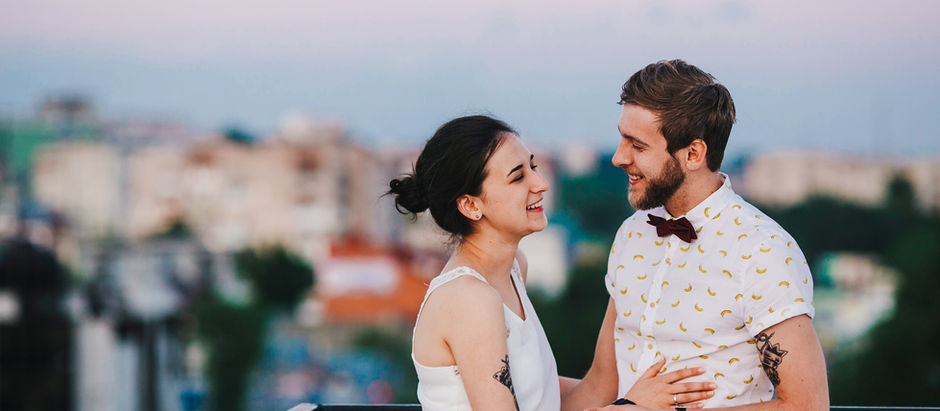 A couple smiles at each other on a rooftop at sunset. The woman wears a white dress, and the man wears a banana-patterned shirt and bow tie.