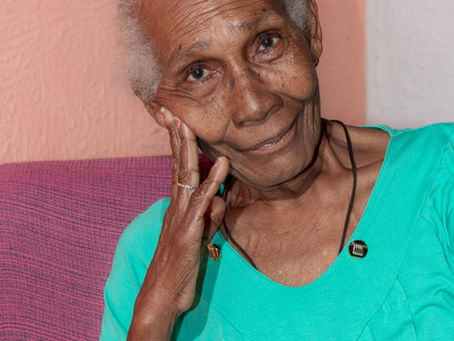 Foto de Dona Catuta, mulher preta de 79 anos. Ela olha para a câmera com a mão direita apoiando a cabeça na bochecha e um sorriso maroto. Ela tem cabelos grisalhos encarapinhados, veste uma blusa verde água e está à frente de uma parede em tom pastel.