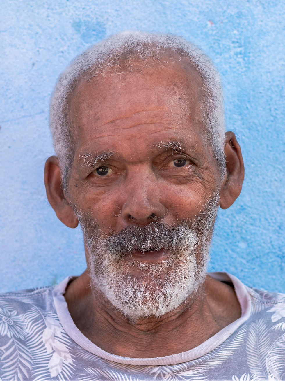 Retrato de Seu Nengo, homem preto idoso. Mostra uma feição serena, apesar de aparentar cansaço. Possui cabelos ralos grisalhos encarapinhados e barba e bigode grisalhos cerrados. Usa uma camiseta estampada e está à frente de uma parede azul clara.