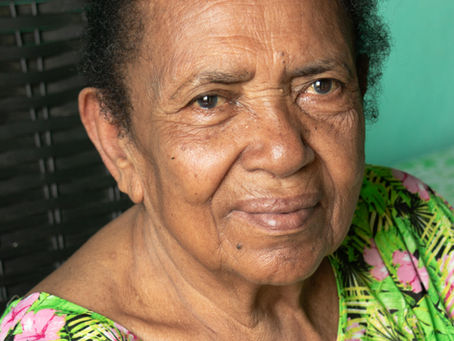 Retrato de Dona Lió, mulher negra idosa, de pele castanha. Ela está sentada, apoiando o queixo sobre a mão direita fechada, em gesto delicado. Tem cabelos presos em um coque, de cor preta. O olhar é sereno e voltado para a câmera, com leve sorriso. Veste uma blusa verde estampada com flores em rosa e amarelo. Ao fundo, uma parede verde e parte de uma cadeira escura, onde ela está sentada.