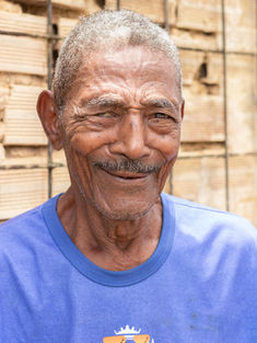 Retrato de Seu Juvenal, homem preto de 88 anos, Apesar da idade, aparenta jovialidade, mesmo com as rugas no rosto. Ele sorri. Tem cabelos curtos grisalhos e encarapinhados, usa bigode fino grisalho. Está vestindo uma camiseta azul e está à frente de uma parede de blocos de cerâmica.