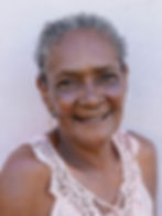 Retrato de Domingas Silva, mulher preta de 73 anos. Ela sorri e forma pequenos vincos entre os olhos, apesar de não ter rugas na testa. Os cabelos são grisalhos e encarapinhados, amarrados para trás. Ela usa uma blusa rosa e brincos com pingente em forma de coração. Ao fundo, uma parede cinza.