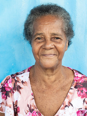Foto mostra o colo e o rosto de uma mulher negra idosa sorrindo, com cabelos encaracolados grisalhos. Ela usa um vestido florido em tons de rosa e está à frente de uma parede azul clara.