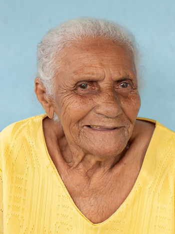 Retrato de Dona Dalva, mulher negra de 91 anos. Ela está com uma feição serena e um sorriso de canto de boca. Ela tem cabelos brancos, veste um vestido amarelo e está à frente de uma parede azul clara.
