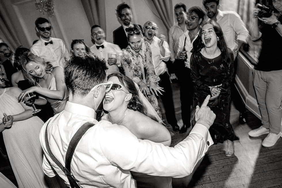 Black-and-white wedding dance floor with a bride and groom kissing as guests in formalwear laugh, sing, and cheer סביב them