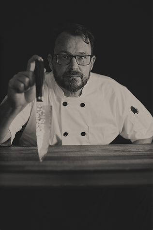 Dramatic black-and-white portrait of a male chef in a crisp white uniform, holding a sharp knife with intensity and focus. The image emphasizes precision, professionalism, and culinary artistry.