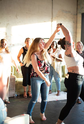 A lively moment at a social gathering, featuring two women dancing together in a sunlit industrial-style venue. A group of smiling attendees watches in the background, adding to the joyful and celebratory atmosphere.