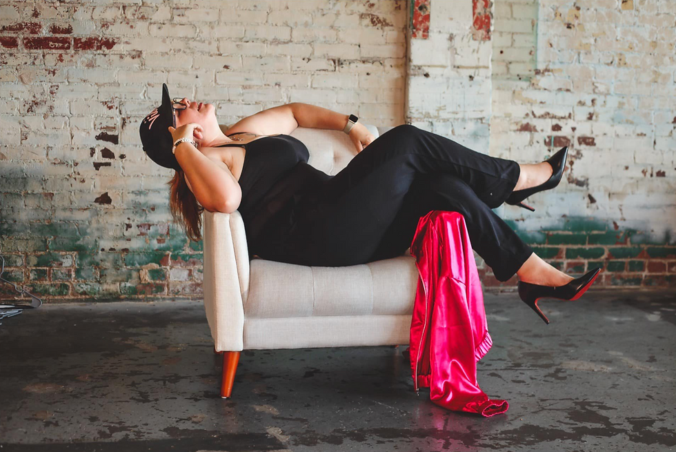 A woman dressed in a black outfit and high heels reclines comfortably on a small white chair in an industrial setting with an exposed brick wall. She wears a baseball cap and glasses, with a hot pink satin jacket draped over the arm of the chair. The setting gives a relaxed and confident vibe, blending casual and elegant elements.