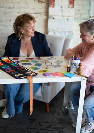 Two women engaged in a creative strategy session at a white table, surrounded by colorful cards, markers, and sticky notes. One woman wears a navy blazer and jeans, while the other, in a pink polka-dot blouse, leans in with a smile. A mug on the table reads 'Live with Intention,' emphasizing a thoughtful and intentional branding approach.