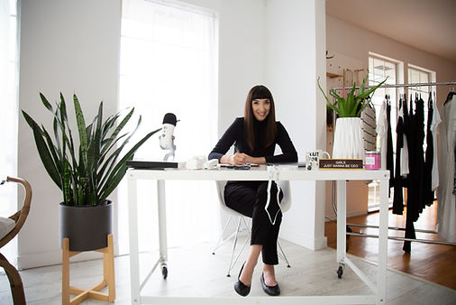 A stylish entrepreneur sits at a bright, modern workspace, surrounded by branding elements that reflect her business. A clothing rack with neutral-toned garments, a microphone, and books with empowering titles add to the aesthetic. Green plants and natural lighting enhance the clean, professional atmosphere, capturing the essence of personal branding photography.