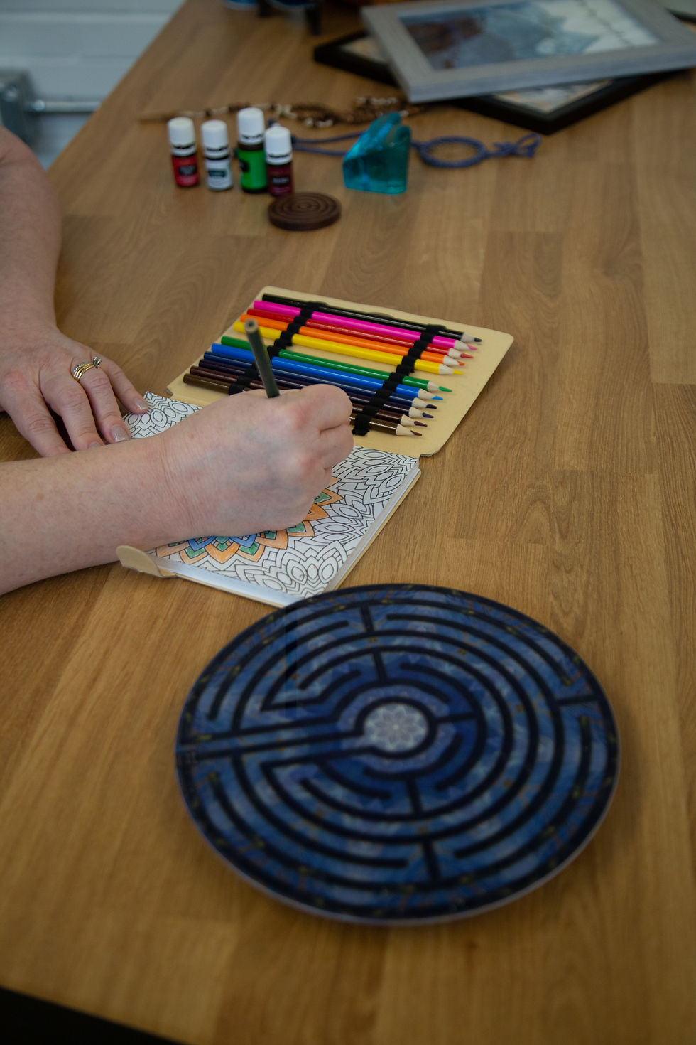 A person is coloring an intricate design on a piece of paper using colored pencils. The pencils are neatly organized in a case, and essential oils and a decorative labyrinth pattern on a blue plate are placed on the wooden table. The atmosphere suggest a calming, creative activity focused on mindfulness or relaxation.
