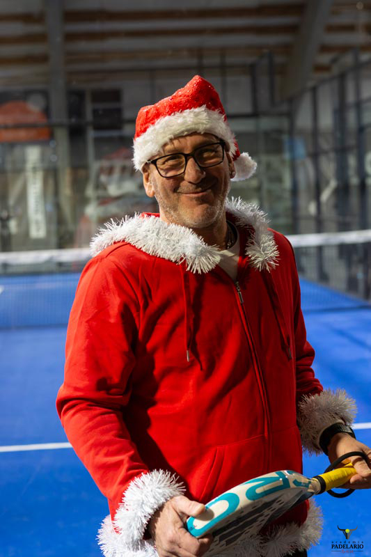 Mann im Weihnachtsmannkostüm, mit Brille und Schläger, auf dem Tennisplatz stehend.