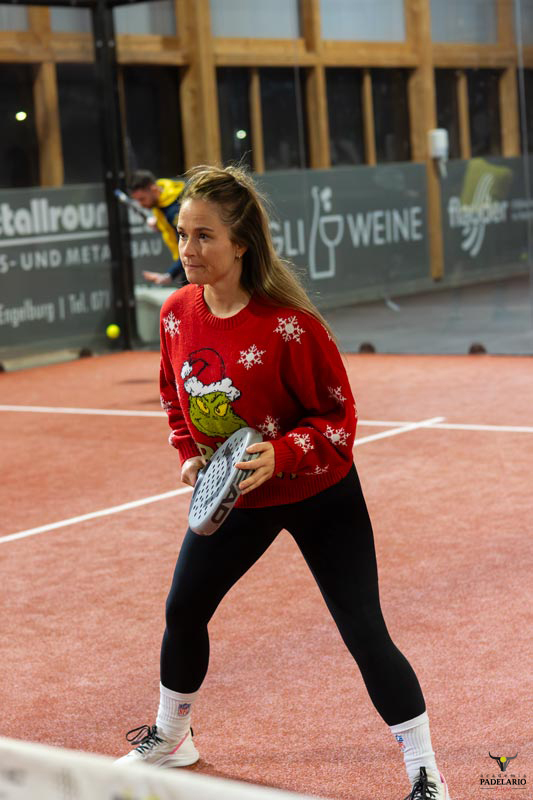 Junge Frau in rotem Pullover spielt Padel, im Hintergrund Werbung auf Deutsch.