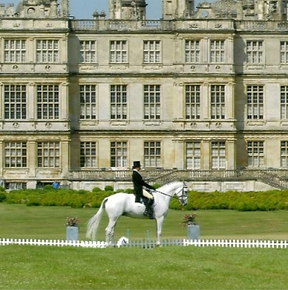 Ginny Howe riding a grey horse in a dressage arna in front of a grand country house