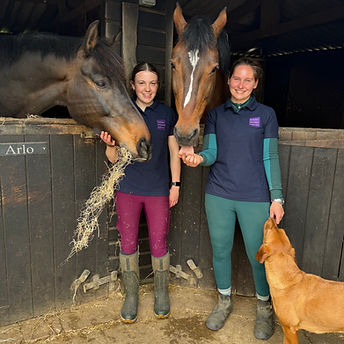 Two women smiling at the camera whilst standing in font of a stable