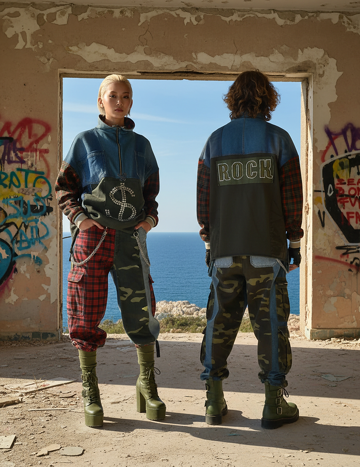 Due modelli posano in una stanza abbandonata con pareti scrostate e graffiti, davanti a una grande finestra che si affaccia sul mare. La modella, di fronte, indossa una giacca in denim con simbolo del dollaro in strass, maniche tartan rosse e pantaloni cargo mimetici con inserti tartan, completati da anfibi verdi con plateau. Il modello, di spalle, indossa una giacca in denim e tessuto militare con la scritta "ROCK" sul retro, pantaloni cargo mimetici e anfibi verdi. Il look esprime lo stile punk, militare e sostenibile della collezione Punk-à-Porter di Marita Balàzs.