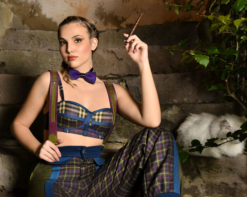 Una modella seduta su una scala, indossando un outfit vintage della collezione Street College, composto da un top a quadri e pantaloni coordinati. Con un'espressione sicura e uno sguardo diretto, tiene un sigaro tra le dita. Il suo papillon viola e i dettagli colorati degli abiti evidenziano il mix audace di stile e eleganza, mentre un gatto bianco riposa accanto a lei, aggiungendo un tocco di calore all'immagine.