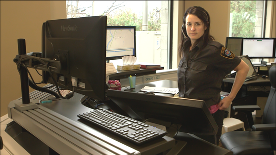 911 dispatcher working comfortably during Telecommunicators Week at a control room workstation, highlighting stacked monitors with both rows of bezels positioned at eye level to prevent neck strain