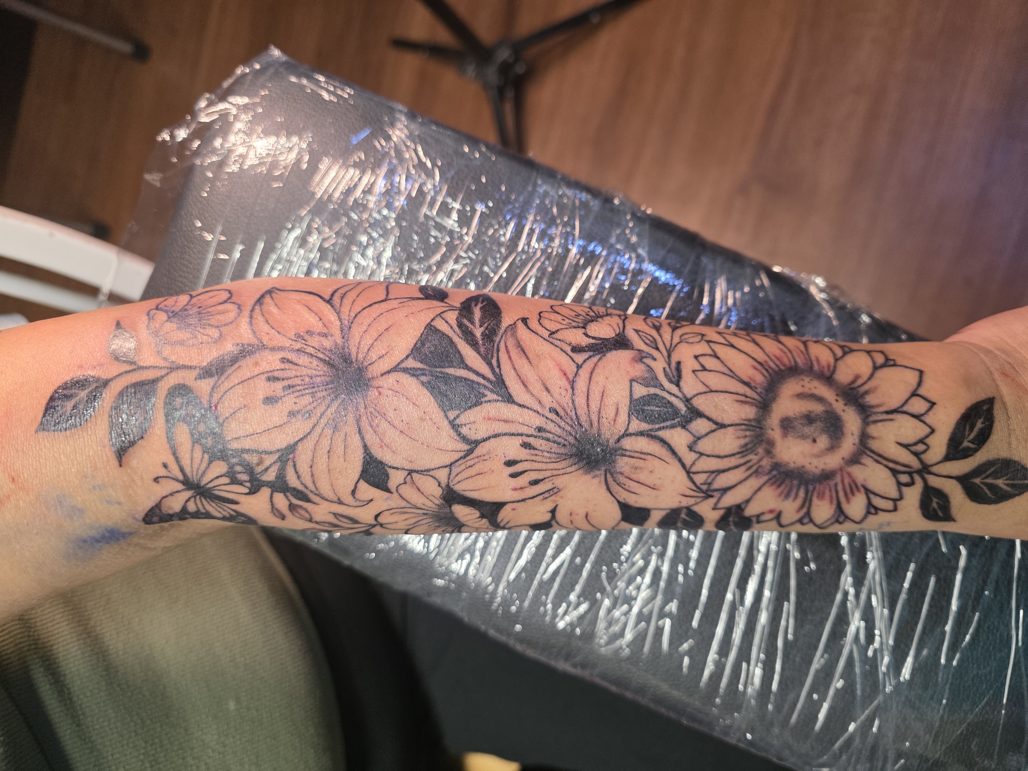 Black line floral tattoo with leaves and a butterfly extending along a forearm; no client face is visible