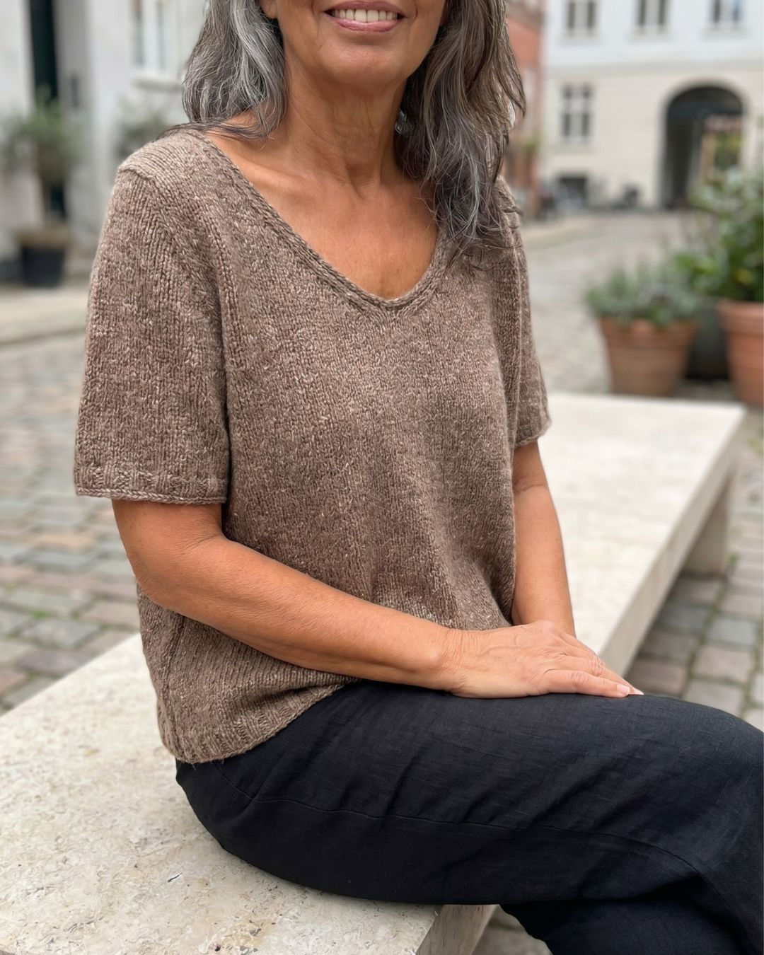 Coco Shirt in braunem Tweedgarn, sitzend auf einer Steinbank getragen, mit lockerem Fall und kurzen Ärmeln.