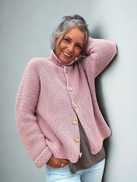Eine lächelnde Frau mit grauem Haar trägt eine handgestrickte, rosafarbene Jacke mit grober Struktur im Perlmuster. Die Jacke ist hochgeschlossen und mit großen Holzknöpfen versehen.