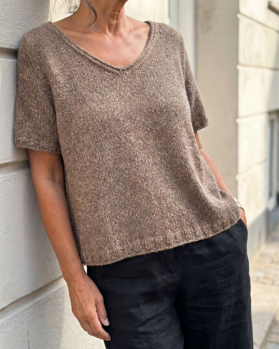 Coco Shirt in braunem Tweedgarn, lässig an einer hellen Hauswand getragen, Ausschnitt, Saum und Maschenbild im Fokus.