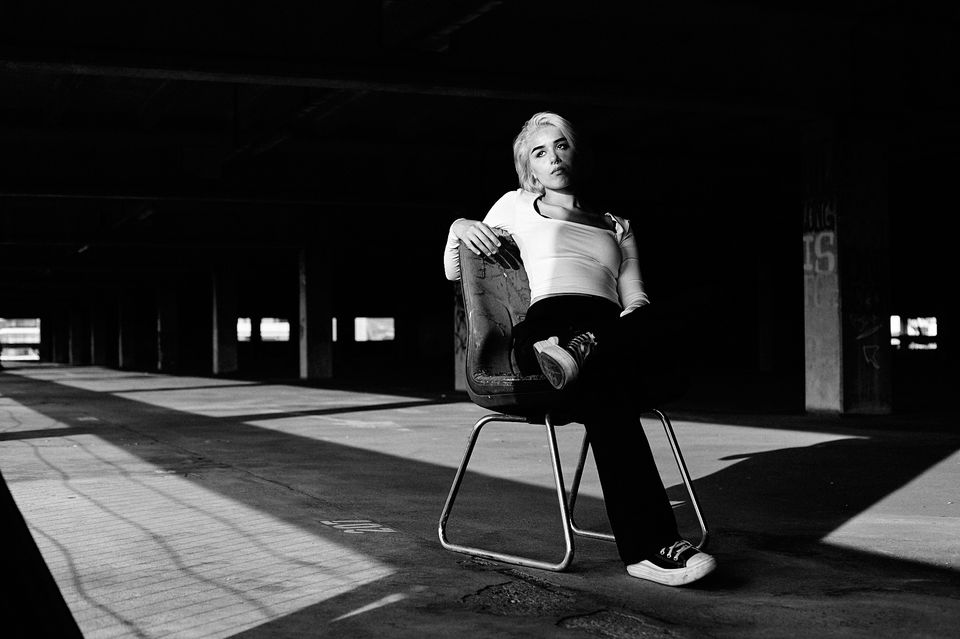 blonde girl sitting on a chair in an empty hall with magic hour, golden hour sun floating the hall. hard shadows, High contrast, low key,  black and white.