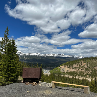Gray small rocks cover the foreground with a simple wooden bench and an old mine cart sit next to one another. The background drops off into a valley lined with pine trees. The background is a the Ten Mile Range with a blue cloudy sky.