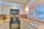 Part of the kitchen includes a range and oven,a microwave, a double sink, cabinets and counters, and a breakfast bar. Beyond the kitchen is shown a bedroom and a wall with photos.