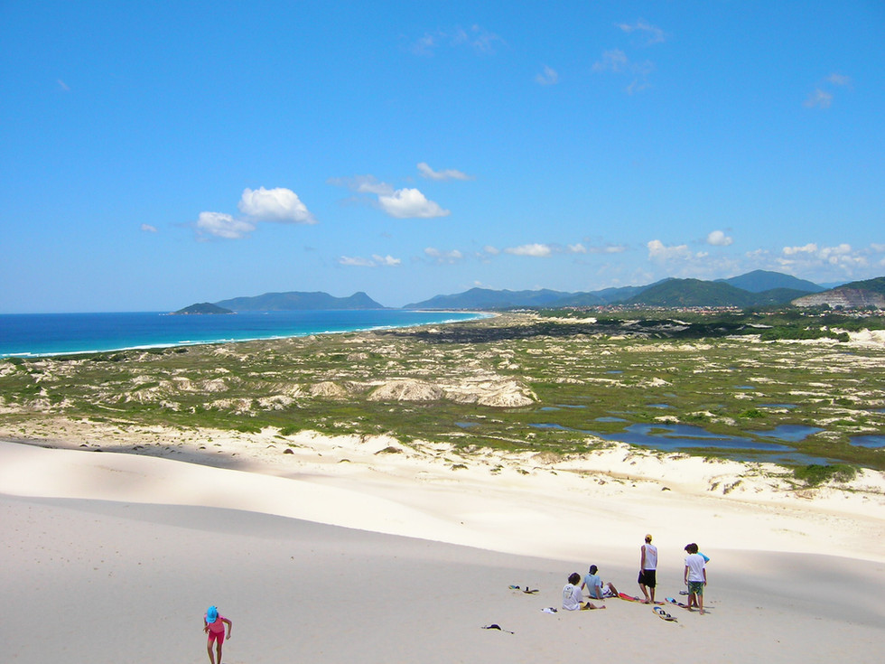 Sob o céu aberto, um grupo descansa, enquanto outro desafia a subida íngreme da duna. A paisagem se abre como um deserto à beira-mar, mas o azul intenso lembra que a vida pulsa logo ali. Entre passos e risos, o vento leva memórias e traz liberdade.
